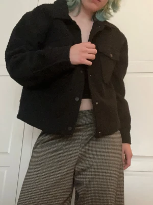 Svart teddy cardiganjacka - Mysig svart cardiganjacka i teddy-material med krage och tryckknappar framtill. Jackan har en bröstficka med lock och är  boxig i modellen. Är 177 för referens. Den är helt oanvänd!