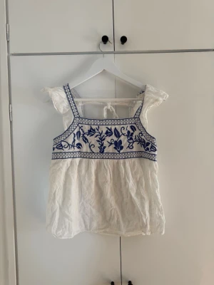 Vit blus med blå broderi och volang - Söt vit blus med blå broderade detaljer och volang vid axlarna. Toppen har breda axelband, fyrkantig ringning och knyt i ryggen. Perfekt för sommarens alla tillfällen och ger en bohemisk vibe.