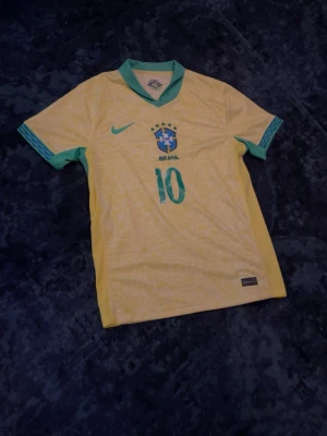 Brasilien Neymar Jr matchtröja  - Säljer en  Brasilien fotbollströja från Nike med Neymar Jr och nummer 10 på ryggen. Tröjan har gröna detaljer vid krage och ärmslut samt Brasilien-emblem på bröstet. Perfekt för dig som älskar fotboll och vill stötta landslaget.
