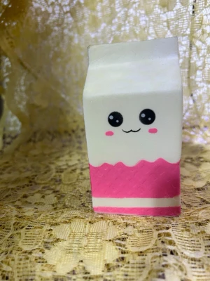 Söt mjölkpaket squishy i rosa och vitt - En squishy i form av ett mjölkpaket med gulligt ansikte på framsidan och ett rosa hjärta på baksidan. Färgerna är vitt och rosa, och materialet är mjukt skumgummi. Perfekt som stressleksak eller dekoration på skrivbordet.