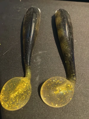 Två gummibeten i svart och guld - Säljer två mjuka gummibeten för fiske, i svart och guldglittrig färg. Betena har paddelformad svans och realistiska ögon, perfekta för predatorfiske. Tillverkade i mjukt plastmaterial som ger naturlig rörelse i vattnet.