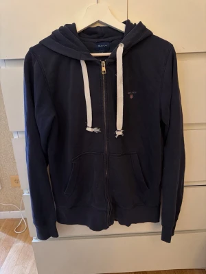 Mörkblå zip-hoodie från GANT - Mörkblå hoodie från GANT med dragkedja, vita grova dragsnören och fickor framtill. Klassisk broderad GANT-logga på bröstet. Mjuk bomullskänsla och huva för extra chill vibe. Perfekt för en avslappnad streetstyle-look. Dustbag medföljer. 
