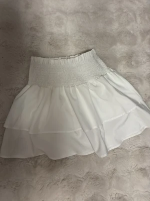 Vit volangkjol med smock - Söt vit kjol med bred smock i midjan och dubbla volanger. Kjolen har en luftig och flowy känsla, perfekt för sommardagar. Materialet ser ut att vara ett tunt och mjukt tyg som ger kjolen ett lätt fall.