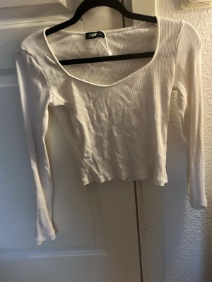 Vit ribbad långärmad topp - Säljer en vit ribbad topp med lång ärm och djup rundad ringning. Toppen är croppad och har en smickrande passform som passar perfekt till jeans eller kjol. Tillverkad i ett mjukt och stretchigt material för extra komfort.