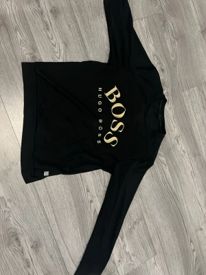 Svart långärmad tröja Hugo Boss - Snygg svart långärmad tröja från Hugo Boss med stort guldfärgat tryck på bröstet. Klassisk rund halsringning och mjukt material som känns skönt mot huden. Perfekt för dig som gillar stilrena och trendiga plagg med tydlig logga.