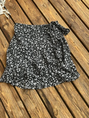 Svart blommig omlottkjol med knyt - Supersöt svart kortkjol med vitt blommönster och omlottdesign. Kjolen har en volangkant och knyts i midjan för design sen har de en dragkedja. Perfekt till sommaren och vårens alla outfits!
