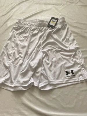 Vita träningsshorts från Under Armour - Säljer ett par vita träningsshorts från Under Armour med elastisk midja och logga i svart på vänster ben. Shortsen är tillverkade i ett lätt och snabbtorkande syntetmaterial som passar perfekt för sport och aktivitet.