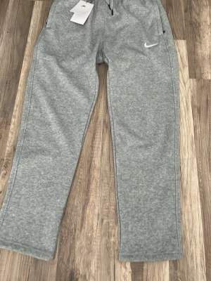 Grå mjukisbyxor från Nike - Snygga grå mjukisbyxor från Nike med klassisk vit logga på benet. Byxorna har dragsko i midjan och sidofickor med dragkedja. Perfekta för chill eller träning, tillverkade i mjukt och bekvämt material.