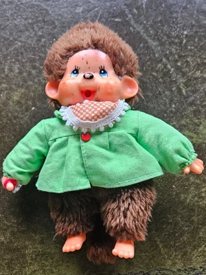 Söt Monchhichi-docka i gröna kläder - En gullig Monchhichi-docka med mjuk, brun päls och gröna tygkläder. Dockan har ett glatt ansikte med blå ögon, rosa kinder och en vit krage med orange mönster. Perfekt för samlare eller som present till någon som gillar retroleksaker.