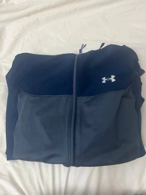  Under Armour - Snygg blå vindjacka från Under Armour med huva och hel dragkedja framtill. Jackan har två sidofickor med dragkedja och reflekterande detaljer på ärmarna. Perfekt för dig som vill ha en sportig look. Den är köpt i Stockholms t central i självaste under armour butiken. Inga tecken på att den är trasig eller i dåligt skick på något sätt har använt den men aldrig veljat använda den så att den blir sliten så den är så gott som ny🤍
