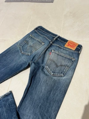 Blå Levi's raka jeans 501 - Klassiska blå Levi's jeans med raka ben och fem fickor. Jeansen har en snygg tvättad look med slitningar och den ikoniska läderlappen bak i midjan. Passar perfekt till en avslappnad stil och är gjorda i slitstarkt denimtyg.
