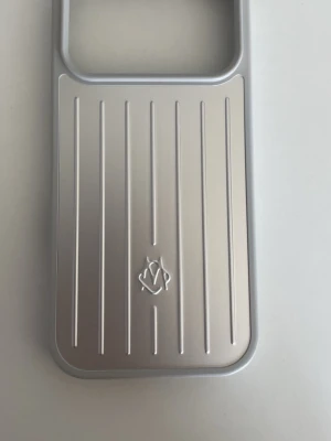 Rimowa IPhone 17 PRO Max skal - Snyggt och stilrent mobilskal i silver från Rimowa. Skalet har en metallisk finish med vertikala ränder och en präglad logotyp på baksidan. Tillverkat i tåligt material och designat för att ge din mobil en exklusiv look.