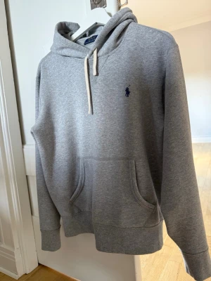 Ralph Lauren Hoodie  - Snygg klassisk grå hoodie från Polo Ralph Lauren med klassisk blå logga broderad på bröstet. Hoodien har huva med vita dragsnören. Storlek på Hoodien är Small och i mycket bra skick. 