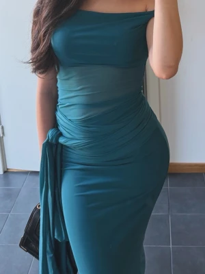 Turkos axelbandslös långklänning - Säljer en elegant turkos axelbandslös långklänning med figurnära passform och draperad midja. Klänningen har en slät yta och ett dekorativt tygparti som hänger ner från midjan. Perfekt för dig som vill sticka ut med en stilren look.