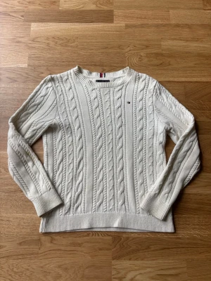 Vit kabelstickad tröja Tommy Hilfiger - Kabelstickad vit tröja från Tommy Hilfiger med rund halsringning och diskret logga på bröstet. Tröjan har långa ärmar och ribbade muddar vid ärmslut och nederkant. Perfekt för dig som gillar klassisk och stilren look.