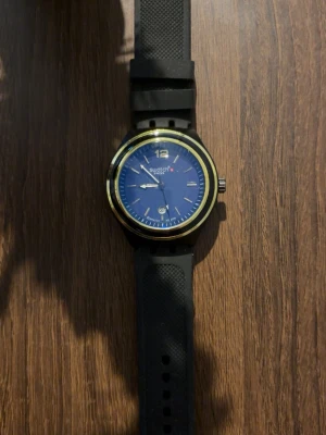Klocka  - Snygg klocka med blå urtavla, svart armband och gulddetaljer runt boetten. Klockan har datumvisning och klassisk rund form. Perfekt accessoar för dig som vill ha en stilren och modern look.