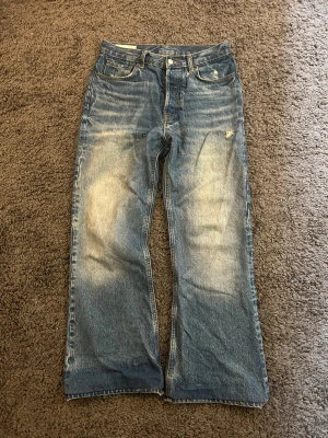 Blåa utsvängda/bootcut jeans med slitningar - Ett par blå bootcut jeans och tydliga slitningar samt ljusa partier framtill och baktill. Jeansen har fem fickor och knappgylf. Materialet ser ut att vara klassisk denim i bomull. Perfekta för en avslappnad och trendig look. Uppsydda fråga om mått 