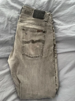 Grå jeans från Nudie Jeans - Säljer ett par grå jeans från Nudie Jeans med klassisk femficksmodell och vågig söm på bakfickan. Jeansen har en avslappnad passform och är tillverkade i mjukt denimtyg. Perfekta för dig som gillar en stilren och enkel look. Notera! De e jätte lite färg stänk på fickan där bak man märker knappt de