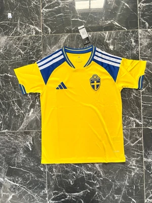 Svensk landslagströja Adidas gul/blå - Gul kortärmad landslagströja från Adidas med blå detaljer och vita ränder på axlarna. Bröstet pryds av Sveriges landslagsemblem och texten 'Sverige' finns i nacken. Perfekt för match eller träning.
