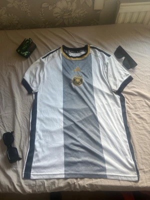 Tysklands matchtröja Adidas - Säljer en officiell Tysklands matchtröja från Adidas med breda vita och grå ränder, gulddetaljer och broderat förbundsmärke på bröstet. Tröjan har korta ärmar, svart/gul krage och är tillverkad i ett lätt och ventilerande material, perfekt för fotboll eller streetwear.Pris kan diskuteras.