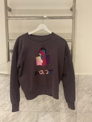 Lila sweatshirt med färgglad logga - Säljer en lila sweatshirt med rund hals och en stor, färgglad Marant-logga på bröstet i olika nyanser av rosa, blått, lila och vitt. Tröjan har ribbade muddar vid ärmslut och nederkant. Perfekt för dig som gillar streetwear och vill sticka ut. Tröjan har två fläckar på framsidan, de går lätt bort i tvätten.