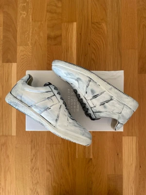 Vitmålade Sneakers Från Maison Margiela - Unika vita sneakers med svartmålade detaljer och svarta skosnören. Skorna har en klassisk låg profil och retroinspirerad design. Tillverkade i läder med slitstark sula. Perfekta för dig som vill sticka ut med en kreativ och edgy look.