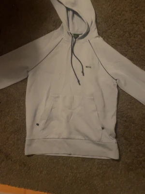 Ljusblå hoodie Hugo Boss - Snygg ljusblå hoodie från BOSS med klassisk huva och dragsnören. Framtill finns en stor magficka och diskret BOSS-logga på bröstet. Hoodien har långa ärmar och ribbade muddar vid ärmslut och nederkant. Perfekt för en chill och avslappnad stil. Använd fåtal gånger