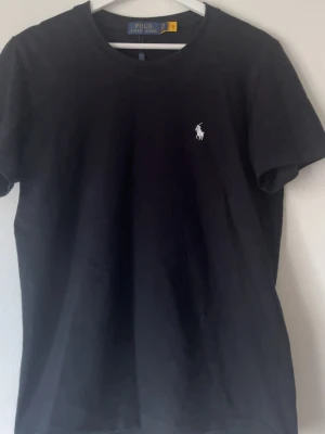 Svart t-shirt från Polo Ralph Lauren - Klassisk svart t-shirt från Polo Ralph Lauren med rund hals och den ikoniska vita logobroderingen på bröstet. Tillverkad i mjuk bomull som känns skön mot huden. Perfekt basplagg för en clean och stilren look.