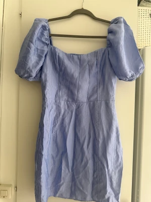 Ljusblå/lila kort klänning med puffärm - Supersöt ljusblå/lila kort klänning med puffiga kortärmar och fyrkantig urringning. Klänningen har en smockad rygg och är tillverkad i ett lätt, tunt tyg som ger en luftig känsla. Storlek 42, väldigt liten i storlek. Upplever den som 36. Kika på de angivna måtten. Dold dragkedja på sidan.
