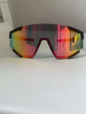 Snabba sportiga solglasögon spegelglas - Säljer ett par sportiga solglasögon från ”inte pr*ada med spegelglas i regnbågsfärger och svart båge. Glasögonen har en futuristisk, kantig form och tydlig logga på sidan. Perfekta för dig som vill sticka ut med en cool accessoar.