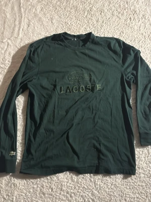 Mörkgrön långärmad topp från Lacoste - Mörkgrön långärmad topp från Lacoste med broderad krokodil och logga framtill. Tillverkad i mjuk bomull och har en klassisk passform. Perfekt för en stilren och avslappnad look.