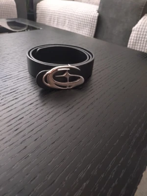 Svart bälte med silverfärgat spänne - Snyggt svart bälte med ett unikt, silverfärgat spänne i metall. Bältet har en stilren och enkel design som passar till många olika outfits. Perfekt accessoar för att lyfta din look.