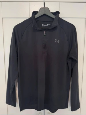 Svart träningströja från Under Armour - Snygg svart långärmad träningströja från Under Armour med halv dragkedja och hög krage. Tillverkad i ett mjukt och stretchigt funktionsmaterial som andas. Perfekt för löpning, gym eller andra sportaktiviteter. Diskret logga på bröstet.