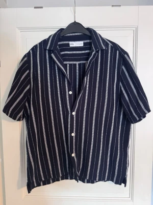 Randig kortärmad skjorta från Zara - Snygg kortärmad skjorta från Zara i marinblått med vita vertikala ränder. Skjortan har klassisk krage, knappar framtill och är tillverkad i ett lätt bomullsmaterial. Perfekt för en avslappnad och trendig look.