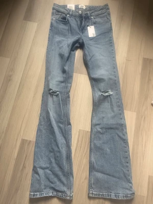 Ljusblå bootcut jeans med hål - Säljer ett par ljusblå bootcut jeans från Lager 157 med slitningar på båda knäna. Jeansen har klassisk femficksmodell, lpg midja och är tillverkade i denim. Perfekta för en avslappnad och trendig look. 