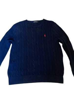 Mörkblå kabelstickad tröja Polo Ralph Lauren - Kabelstickad mörkblå tröja från Polo Ralph Lauren med rund halsringning och den klassiska röda logotypen broderad på bröstet. Tröjan har långa ärmar och ribbade muddar vid ärmslut och nederkant. Perfekt för en stilren och avslappnad look. Mycket bra skick. Storlek S. Se måtten på sista bilderna