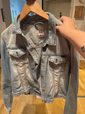 Ljusblå jeansjacka från Gina Tricot - Snygg ljusblå jeansjacka från Gina Tricot med klassisk krage, bröstfickor med lock och knappar framtill. Jackan har en avslappnad passform och coola slitningar för en trendig look. Perfekt att slänga över en hoodie eller t-shirt.
