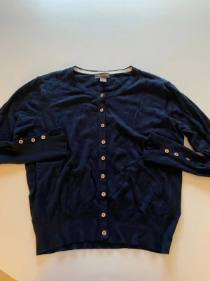 Mörkblå kofta från Primark - Stilren mörkblå kofta från Primark med rund halsringning och vita knappar framtill. Långärmad modell med ribbade muddar och knappar även vid ärmsluten. Perfekt att slänga över en t-shirt eller linne för en enkel och snygg look.