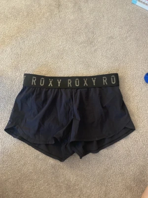 Svarta träningsshorts från Roxy - Snygga svarta shorts från Roxy med bred resår i midjan där ROXY-loggan upprepas runtom. Perfekta för träning eller chill, med en loose passform och sportig look.