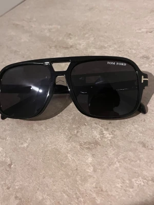 Svarta Tom Ford solglasögon - Snygga svarta solglasögon från Tom Ford med bred båge och fyrkantig form. Klassisk logga på sidan och diskret gulddetalj vid gångjärnen. Perfekta för dig som vill ha en stilren och trendig look.