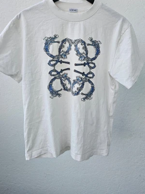 Vit Loewe t-shirt med blått tryck - Snygg vit t-shirt från Loewe med ett unikt blått och svart grafiskt tryck på bröstet. Klassisk rund halsringning och korta ärmar. Perfekt för dig som gillar stilrena plagg med en twist. Tillverkad i mjuk bomull för skön känsla.