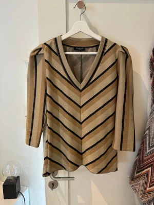 Randig glittrig topp från Selected Femme - Snygg topp från Selected Femme med glittriga guld- och svarta ränder i ett chevronmönster. V-ringad med trekvartsärm och lätt puff vid axlarna. 