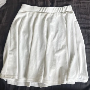 Vit kortkjol i mjukt material - Snygg vit kortkjol med enkel design och lätt utsvängd form. Kjolen har bred linning och är tillverkad i ett mjukt, stretchigt material som ger en bekväm passform. Perfekt att styla med en crop top eller hoodie.