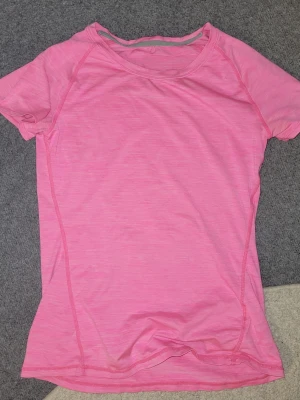 Rosa sportig t-shirt i funktionsmaterial - Snygg rosa t-shirt i funktionsmaterial, perfekt för träning. T-shirten har korta ärmar och en klassisk rund halsringning. Materialet är lätt och andas, vilket gör den bekväm att bära under aktiva dagar.