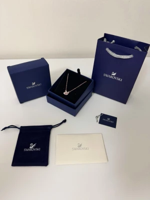Rosa swarovski halsband  - Elegant halsband från Swarovski med ett svanformat hänge dekorerat med gnistrande vita och rosa kristaller. Kedjan är tunn och guldfärgad. Kommer med originalförpackning, presentpåse och certifikat. Perfekt för dig som gillar stilrena och glittriga accessoarer.