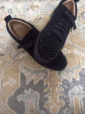 Svarta Louboutin sneakers med nitar - Snygga svarta sneakers från Christian Louboutin med mattsvarta nitar på tån och klassisk röd sula. Skorna har snörning, låg profil och är tillverkade i mocka och läder. Perfekta för dig som vill sticka ut med en edgy och exklusiv look.