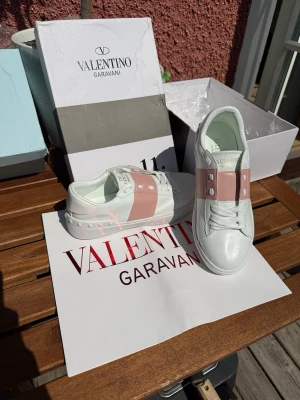Valentino Garavani vita sneakers - Snygga vita sneakers från Valentino Garavani med bred rosa rem över vristen och silverfärgade nitar. Skorna är låga och har en chunky sula samt detaljerad häl. Tillverkade i slätt läder för en clean och exklusiv look.