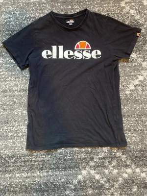 Svart Ellesse t-shirt med logga - Svart t-shirt från Ellesse med stor vit logga och färgglad detalj på bröstet. Klassisk rund hals och korta ärmar. Tillverkad i mjuk bomull för en bekväm känsla. Perfekt för en avslappnad och sportig stil. Storlek 158-164, mycket bra skick