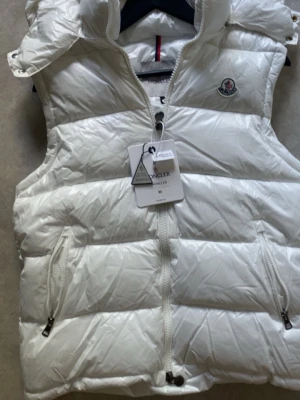 Vit dunväst från Moncler - Snygg vit dunväst från Moncler med hög krage och dragkedja framtill. Västen har två fickor med dragkedja och ikonisk Moncler-logga på bröstet. Insidan har ett färgglatt serietryck och justerbar dragsko nedtill. Perfekt för lager-på-lager och streetstyle.tyvärr kunde jag inte ta så bra bilder för min mobil e trasig😅