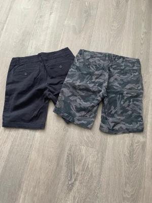 Två par shorts från cubus, marinblå & camo - Säljer två par shorts från cubus, ett par i marinblått och ett par med grått camouflage-mönster. Båda har klassisk passform, sidofickor och bakfickor med knapp. Perfekta för sommaren och tillverkade i mjuk bomull.